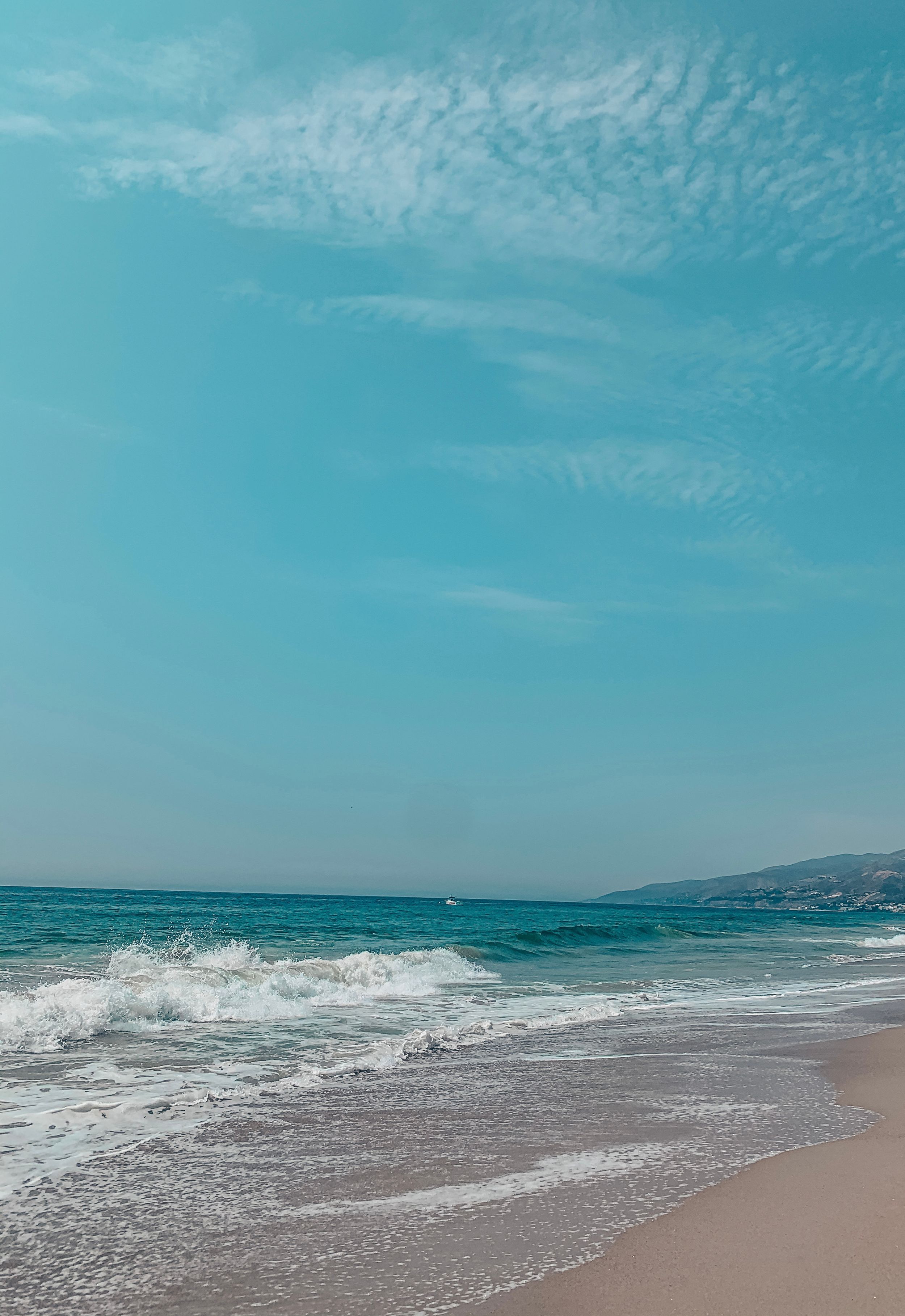 Ocean waves crash onto a sandy beach under a bright turquoise sky with wispy clouds and distant mountains.