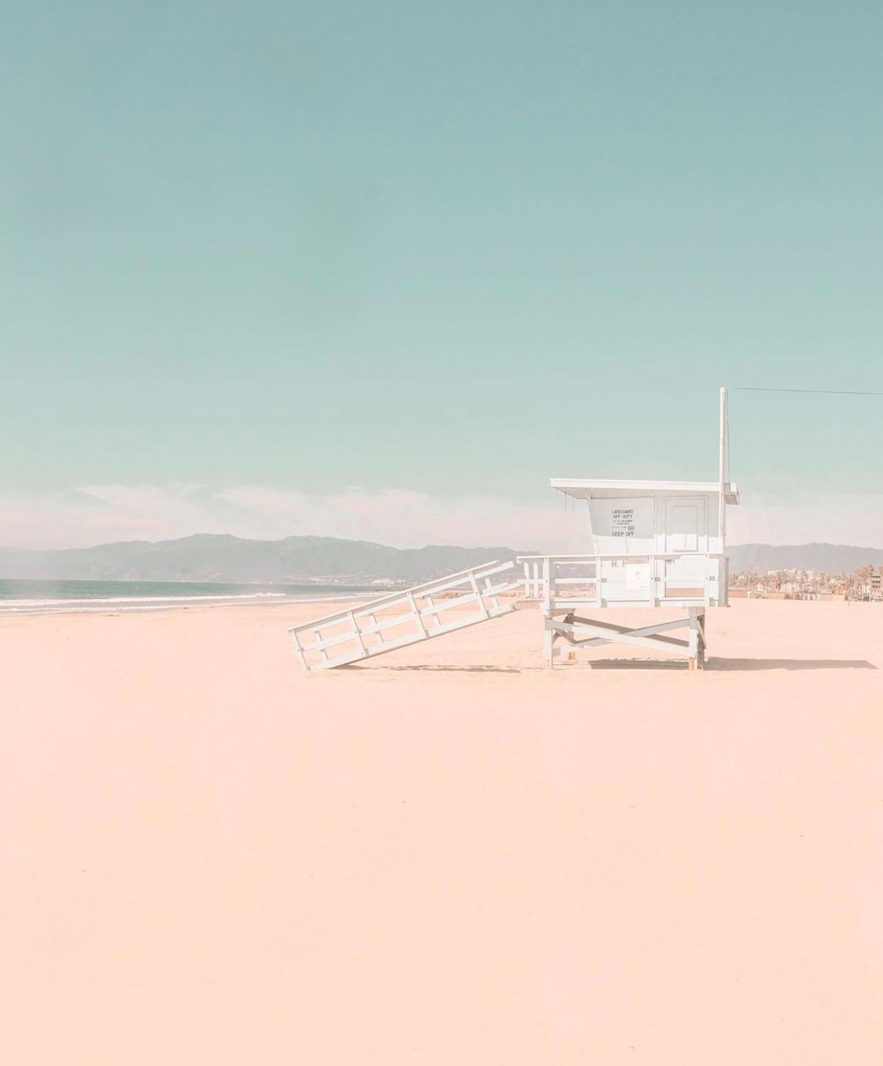 A white lifeguard tower stands on an empty beach with mountains in the distance under a pale blue sky.