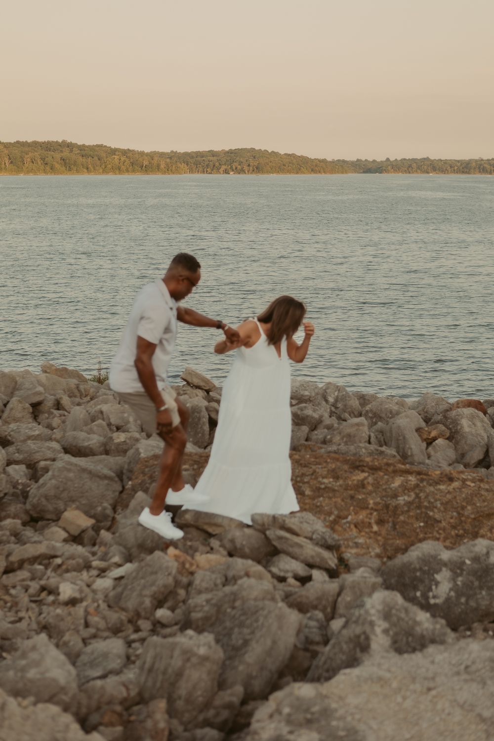 A couple in white attire sits together on coastal rocks overlooking a serene lake at sunset.