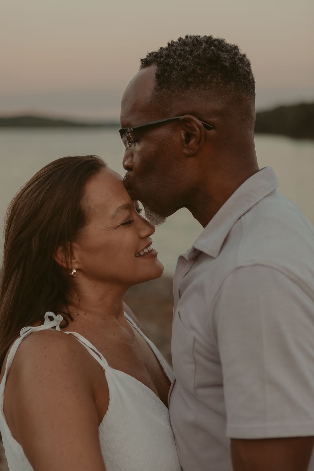 A romantic couple embraces at sunset by a lake, sharing an intimate moment together in warm evening light.