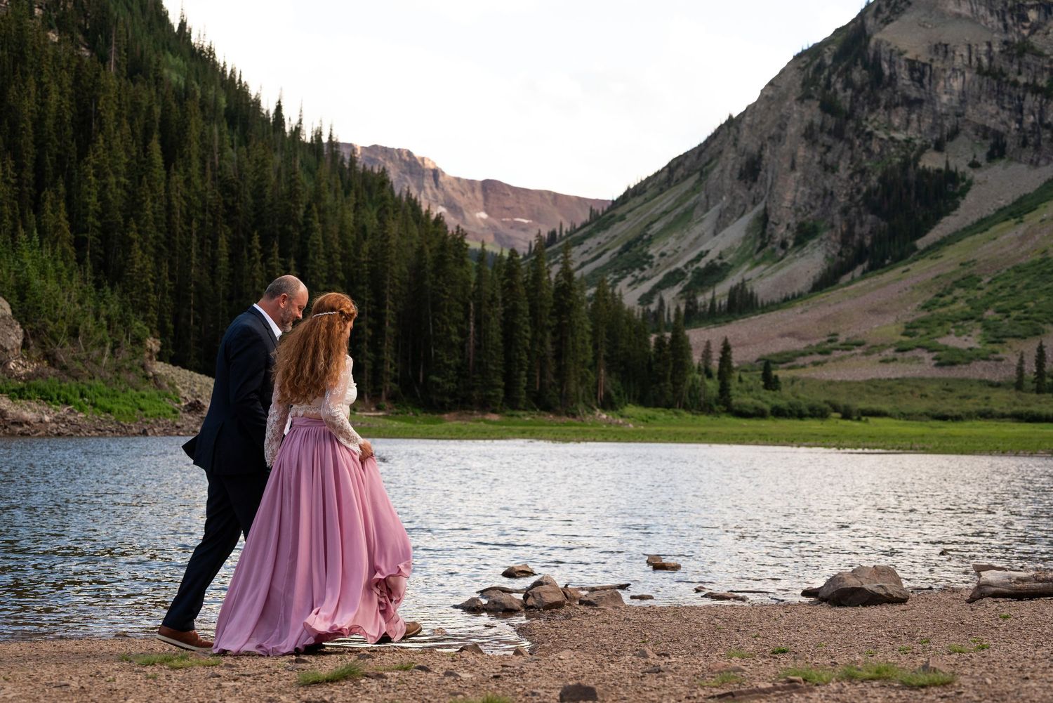 Scenic mountain lake engagement photo with couple standing on rocky shore.