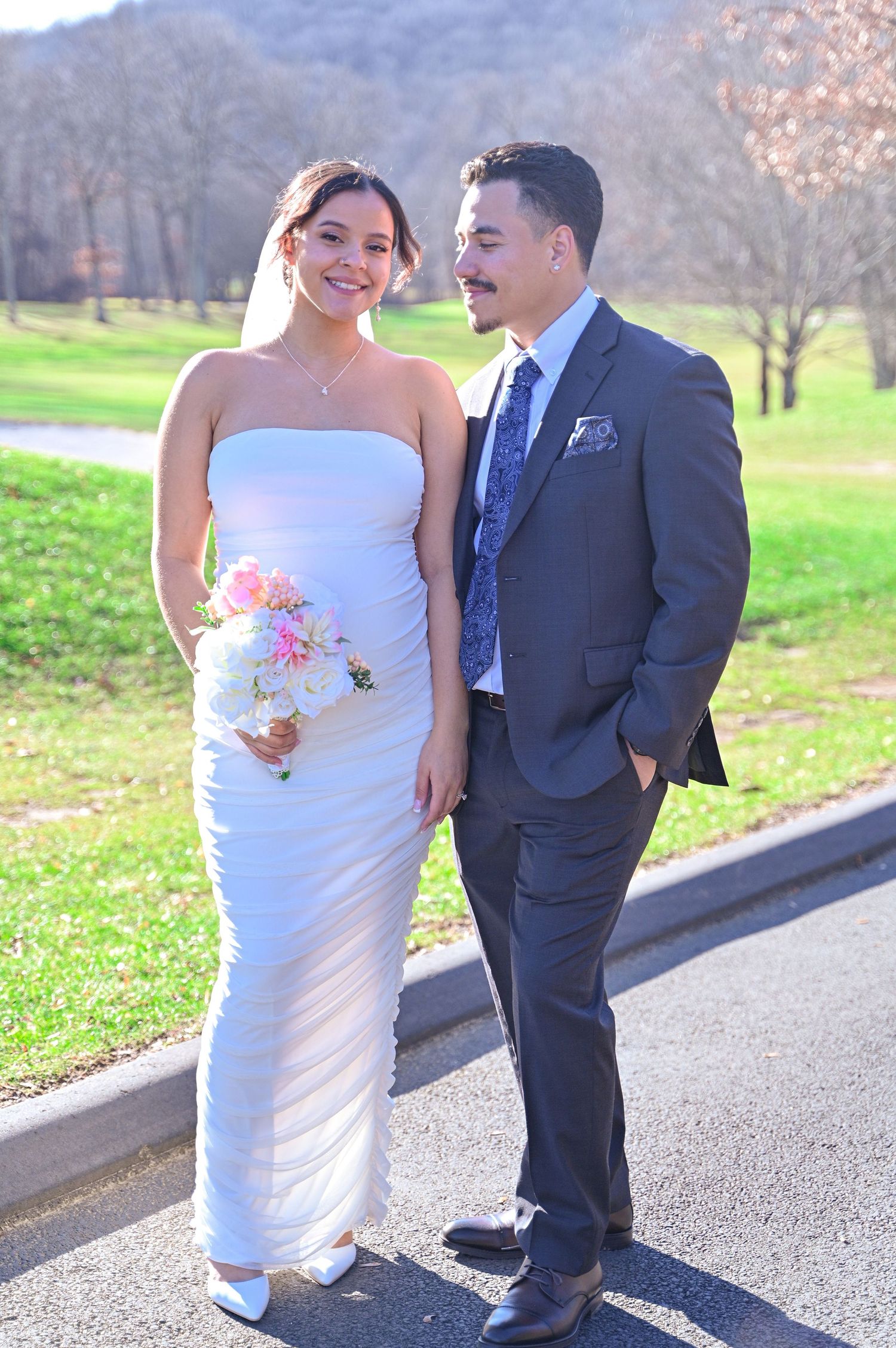 A couple poses outdoors on a sunny spring day with mountains and green grass in the background.