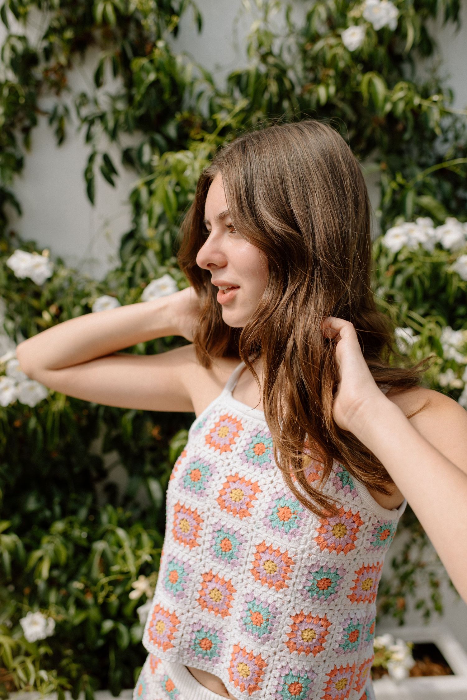 A person stands in front of leafy white flowering vines wearing a patterned crochet tank top on a sunny day.