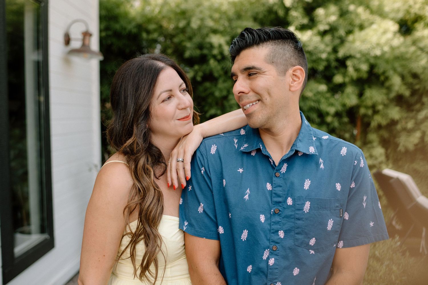 A joyful couple shares a candid moment together outdoors, with a blue patterned shirt and white top against greenery.
