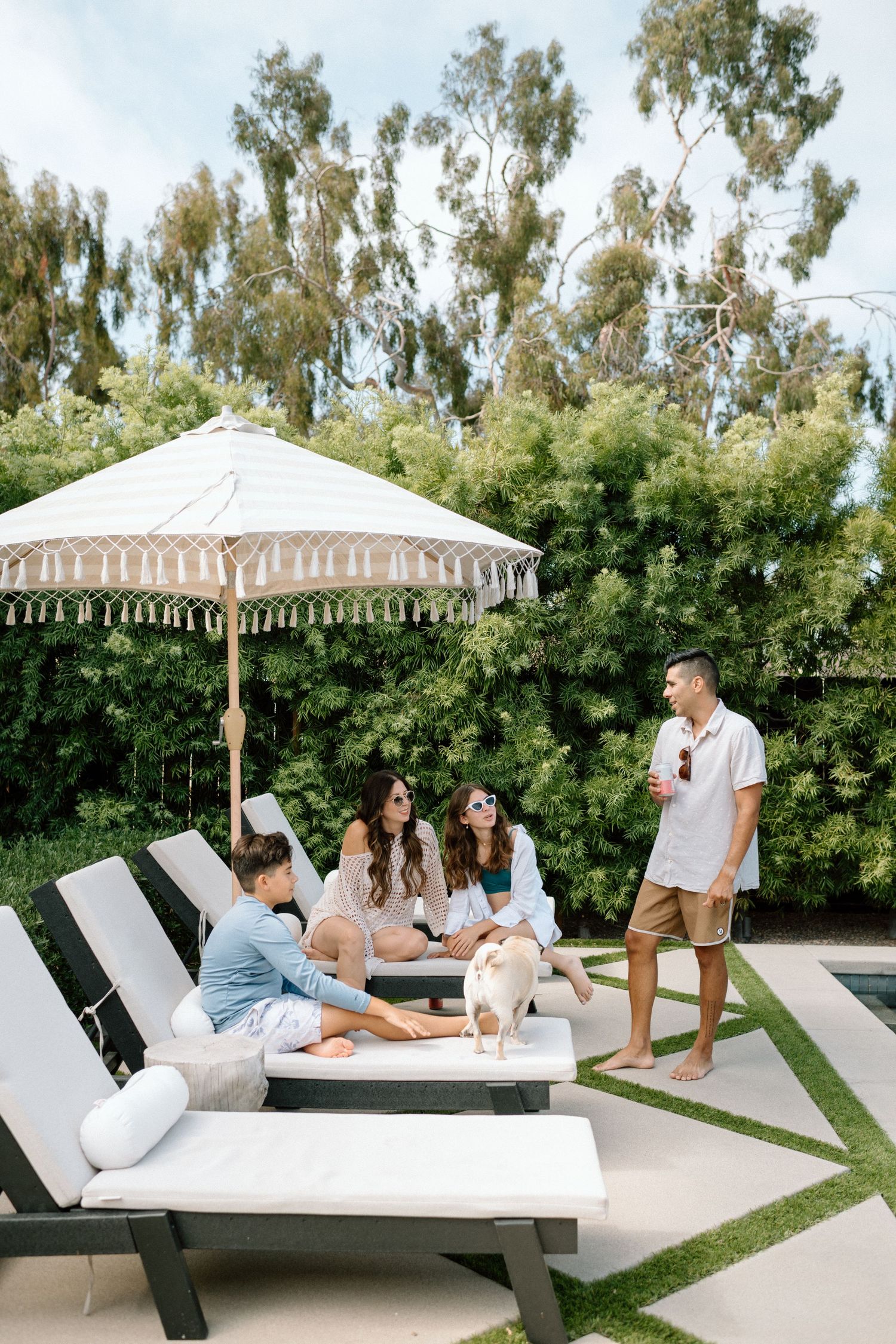 People relaxing on modern white lounge chairs beneath an umbrella in a landscaped backyard oasis.