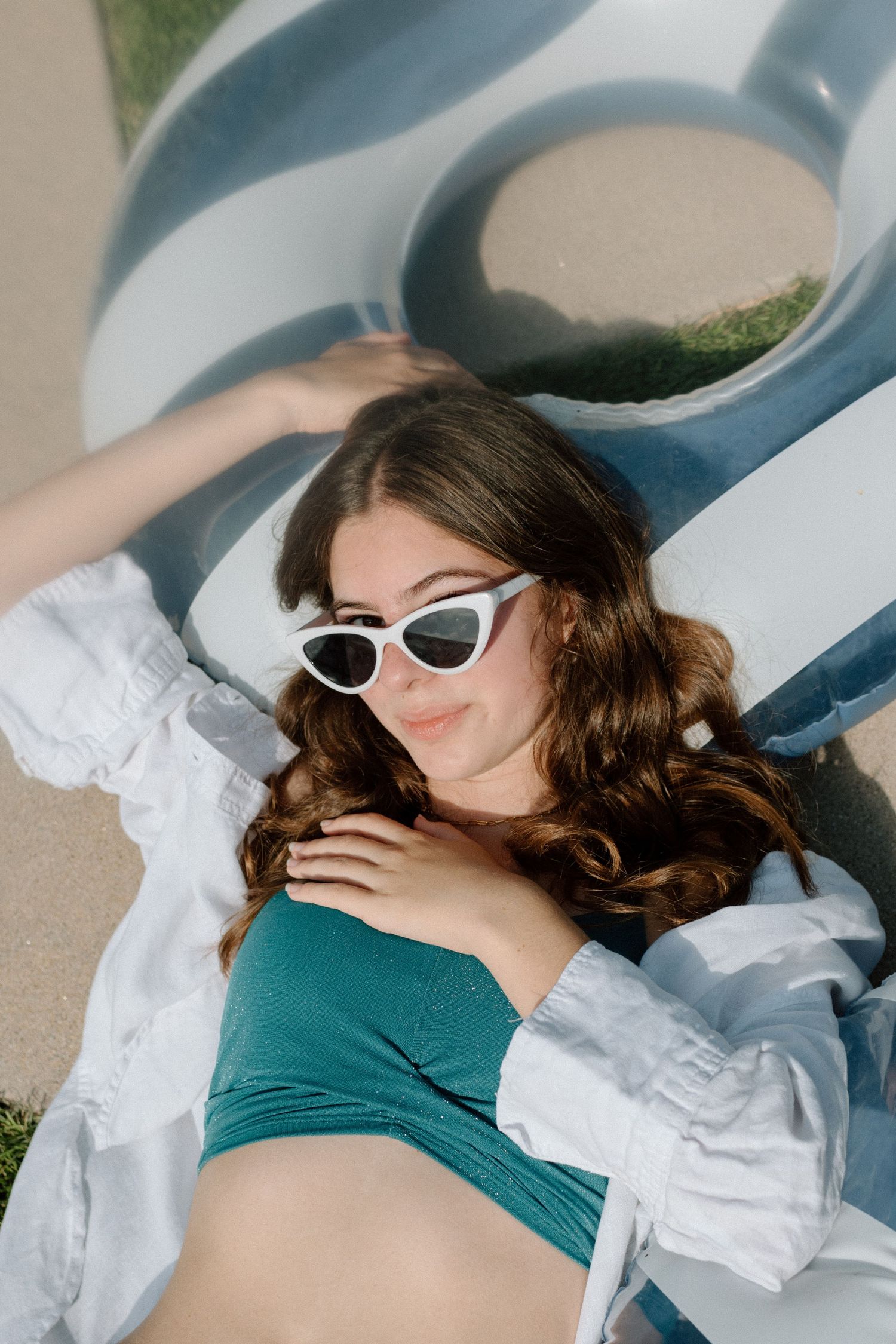 Person in white cat-eye sunglasses and teal swimsuit lying on pool float in summer sunshine.