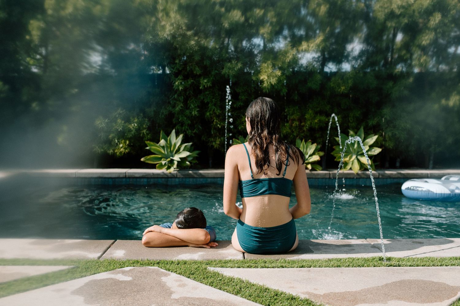 People relaxing by a serene swimming pool surrounded by lush greenery on a sunny summer day.