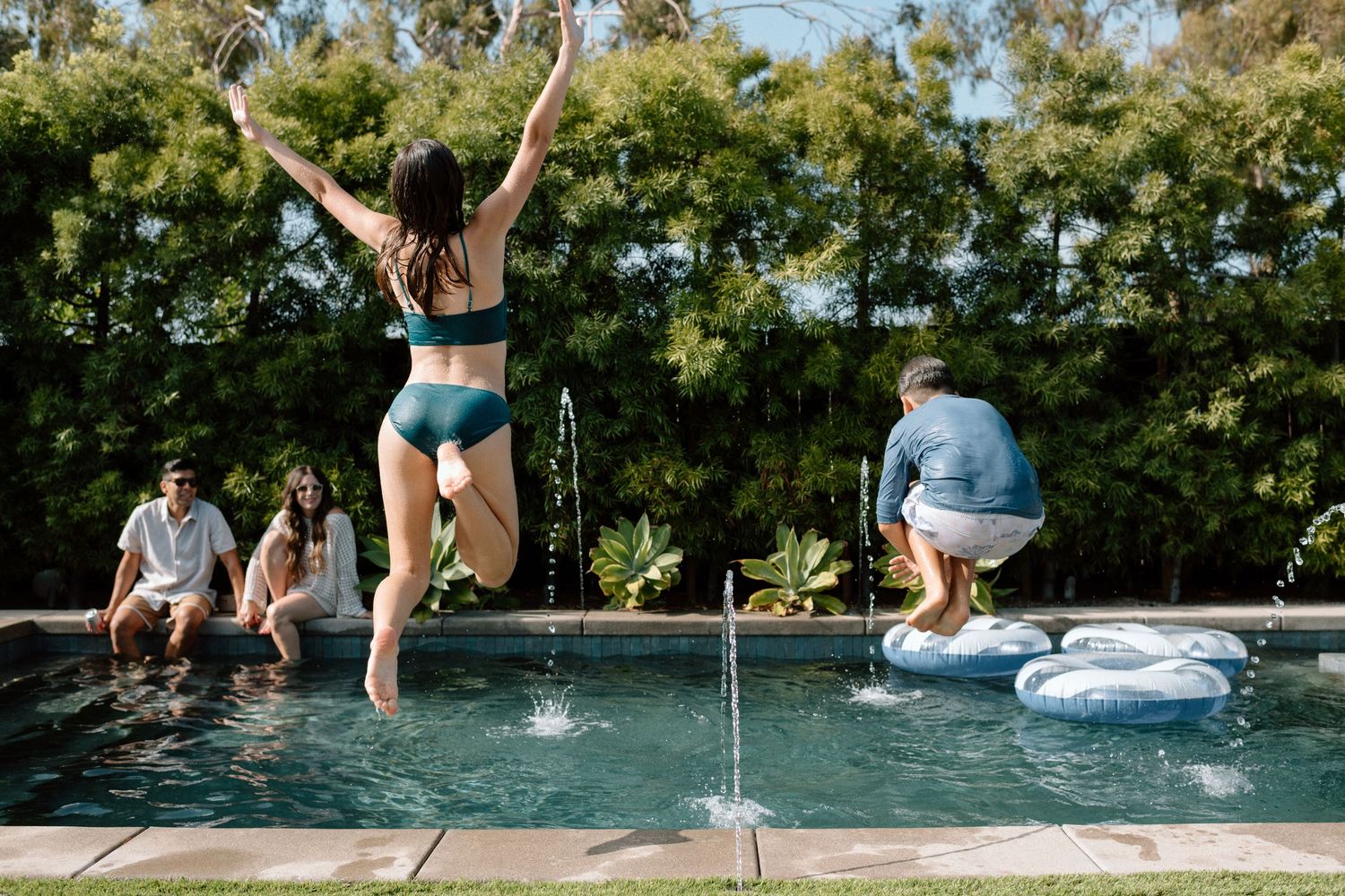 Young people having summer fun jumping into a backyard swimming pool on a sunny day while others watch from poolside.