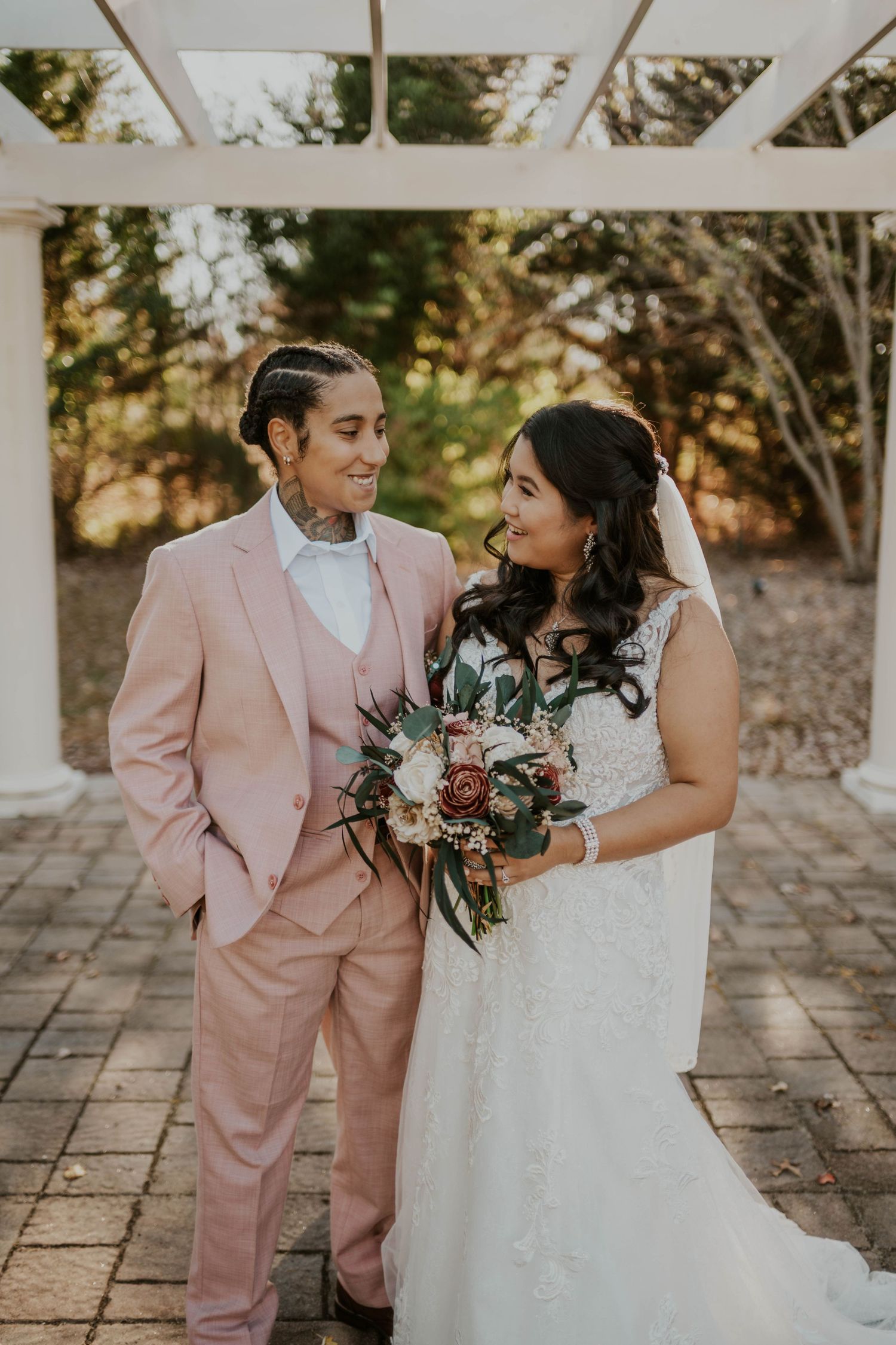 Wedding couple in pink suit and white dress share intimate moment under pergola.