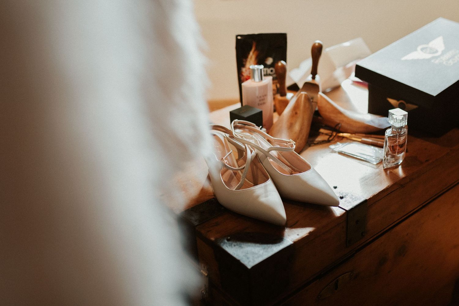 White bridal shoes and jewelry laid out on wooden surface with perfume bottles and accessories in warm lighting.