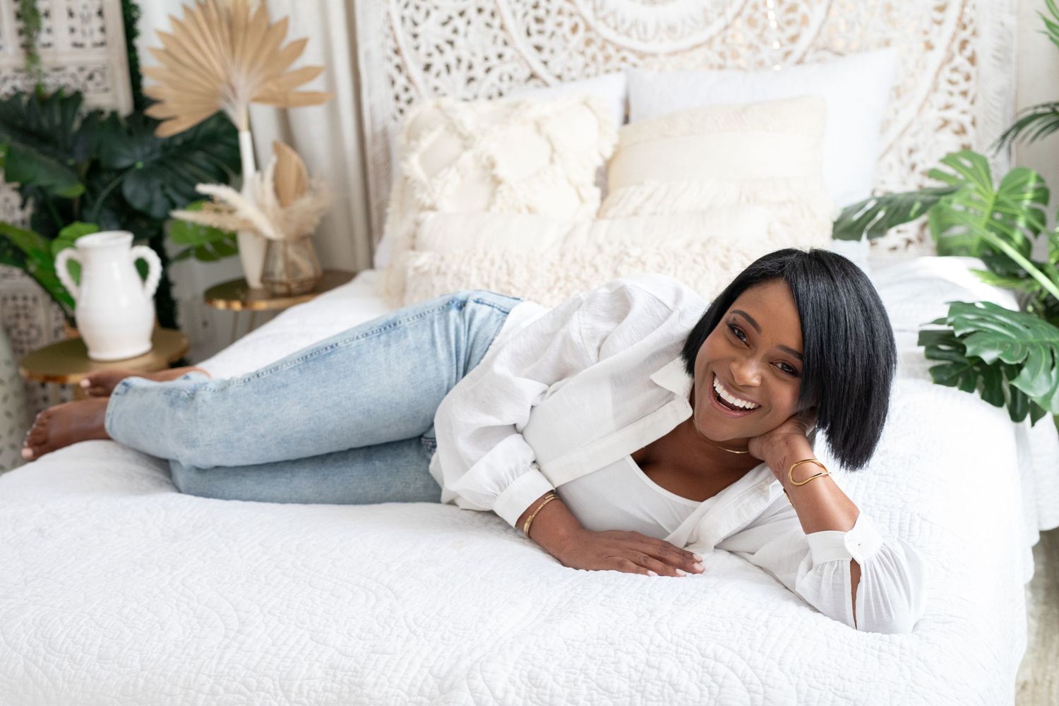 A relaxed individual lounges on a white bed with decorative pillows, surrounded by plants and boho decor.