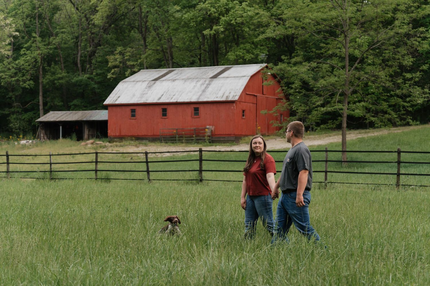 A romantic couple embrace in front of a red barn on a rustic farm with green trees in the background.