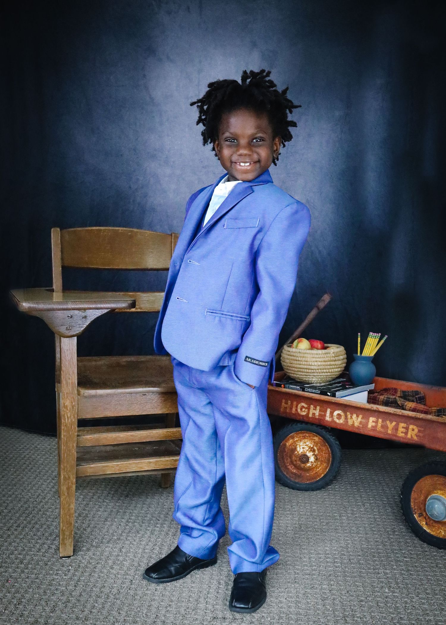 A bright-eyed youth in a light blue suit poses beside a vintage wooden chair and red Radio Flyer wagon with school supplies.