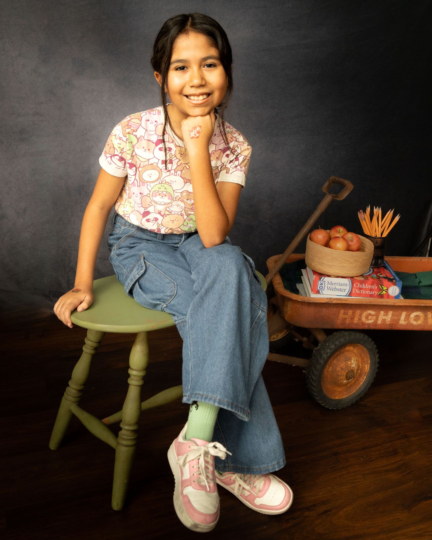 A young person in a floral shirt and jeans sits on a green stool with a vintage red wagon prop in the background.