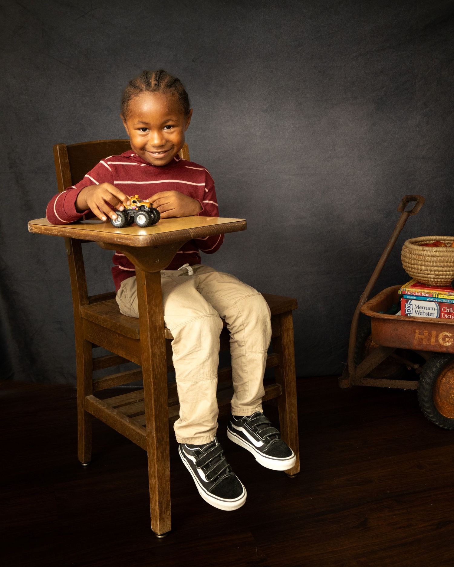 Photography of a child sitting in a wooden chair wearing burgundy shirt and beige pants with toy camera in a dark studio setting.