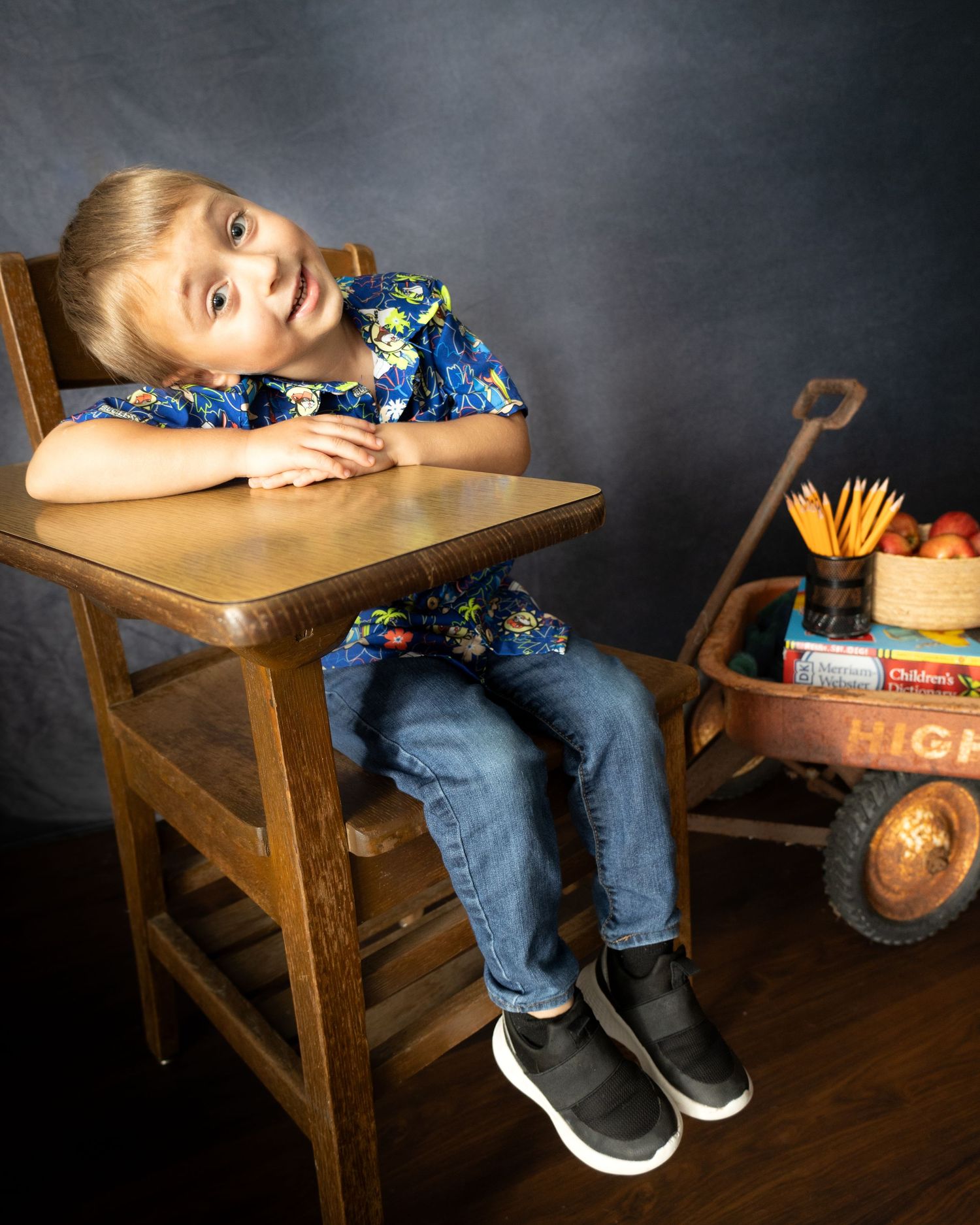 Five sequential shots show a small child in a blue shirt and jeans playfully draped over a wooden school desk against dark backdrop.