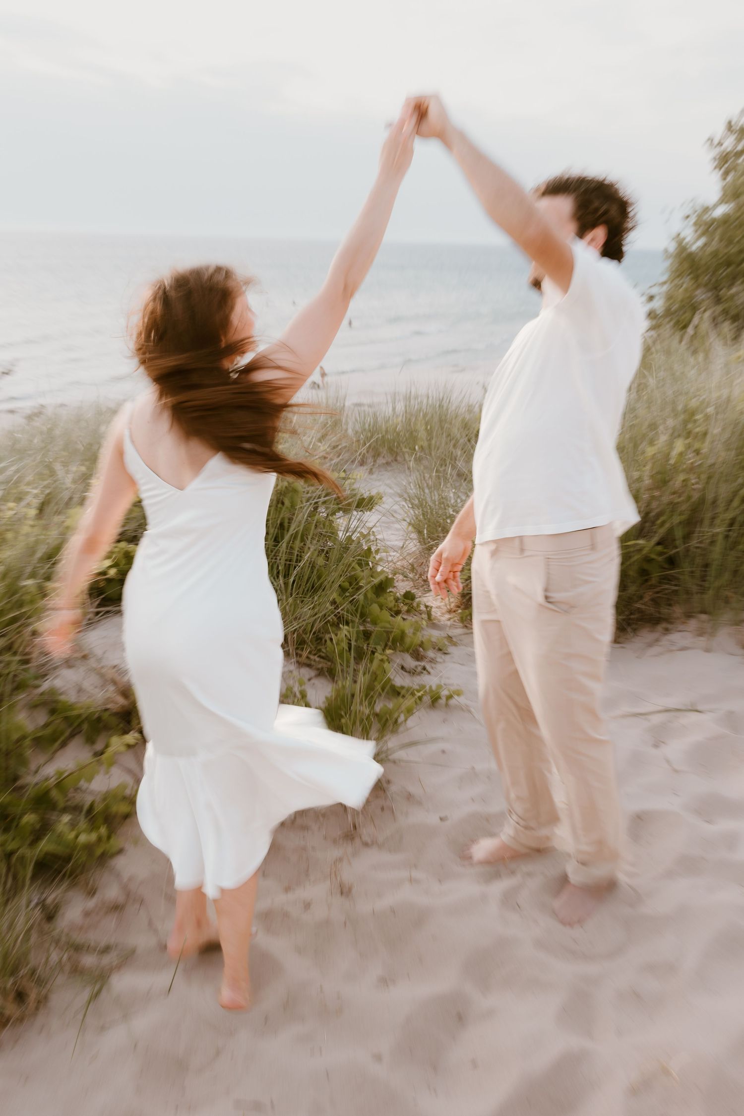 Two people dancing joyfully on a sandy beach at sunset, their bodies creating flowing movements in white and beige outfits. Michigan Wedding
