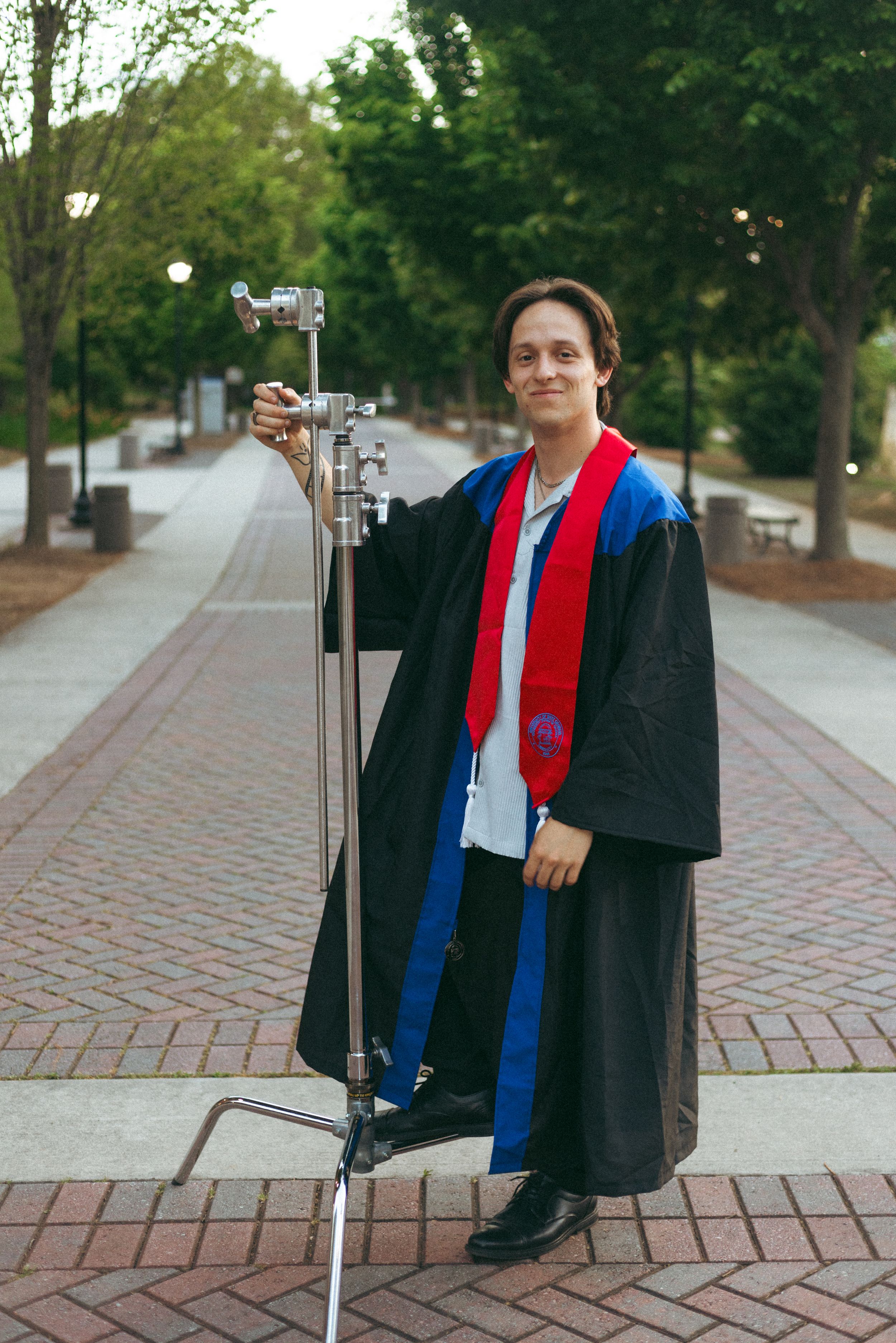 A graduate in academic regalia poses with a c-stand on a brick path surrounded by trees on a university campus.