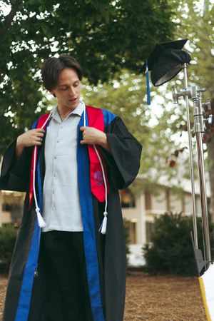 A graduate wearing their academic regalia adjusts their cap and gown while preparing for commencement.