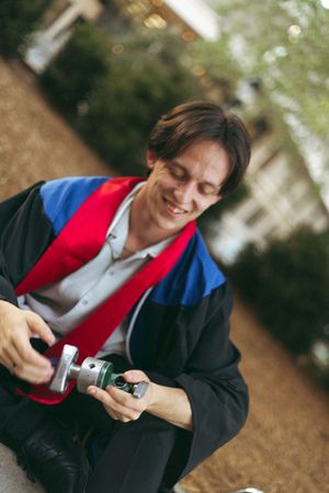 A figure wearing a black cardigan and red vest sits outside while examining a small object in their hands.