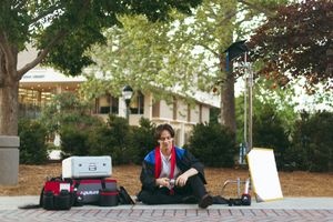 A DJ sets up their equipment outdoors with speakers and gear on a patio under green trees.