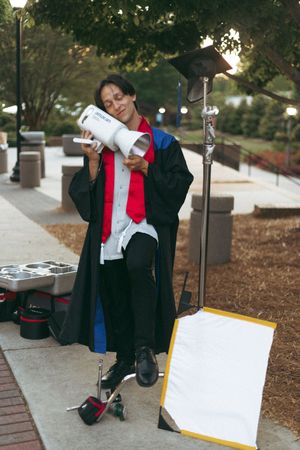 A graduate wearing traditional black academic regalia with red accents sits and poses for photos outdoors.
