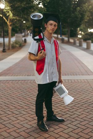 Graduate in cap and gown holding megaphones stands on brick pathway surrounded by trees.
