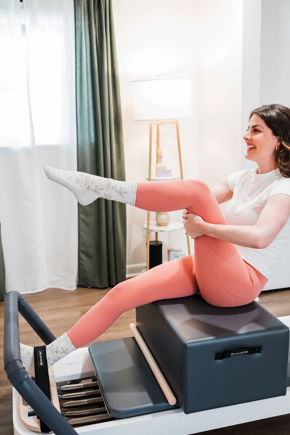 Person demonstrating Pilates exercises on black reformer machine in bright home studio.