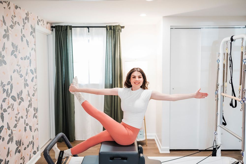 Person performing a joyful pose on pilates equipment while wearing coral leggings and white top.