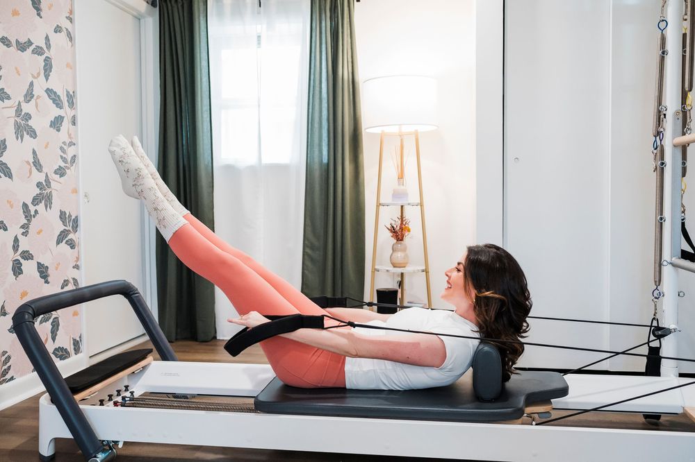 Person exercises on a pilates reformer machine in a bright room with green curtains and floral wallpaper.