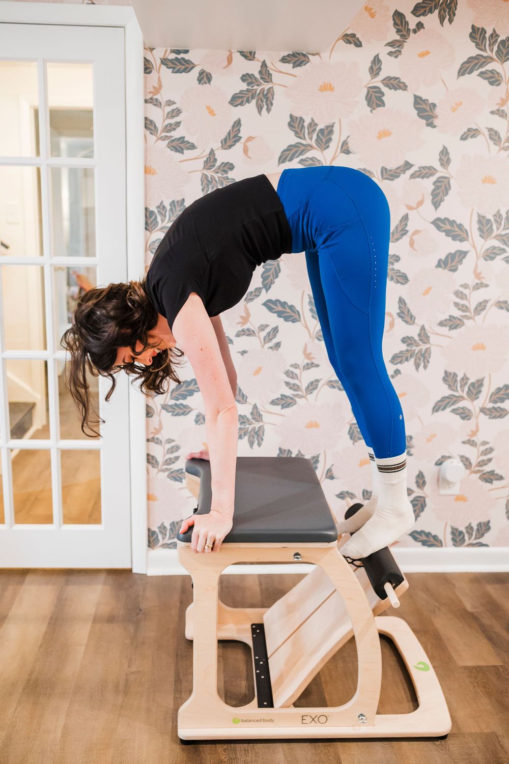 Demonstrating proper form for exercise movements using wooden Pilates equipment against floral wallpaper.