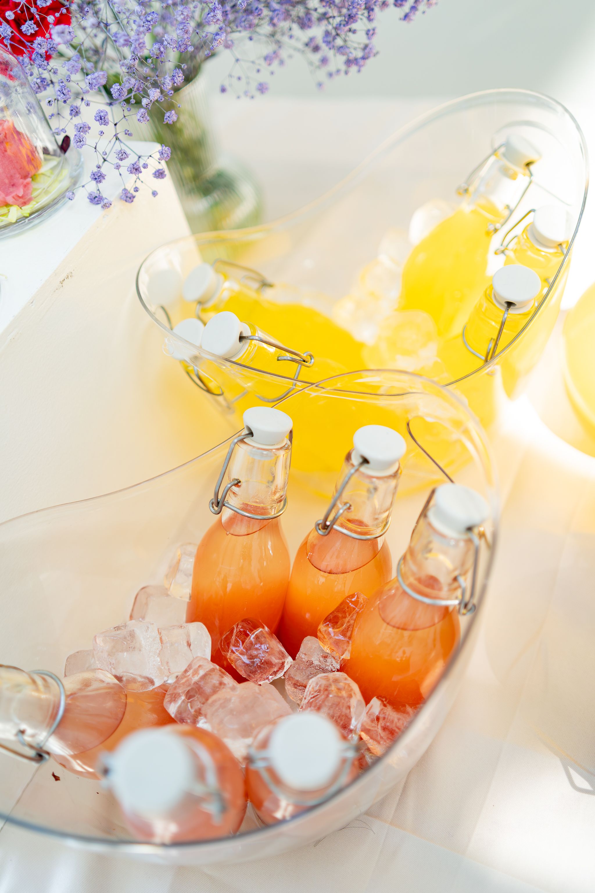 Glass bottles of orange and yellow beverages chill on ice with purple flowers for decoration.