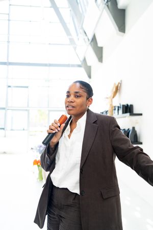 A person wearing a brown blazer and white shirt gestures while speaking in a bright room.
