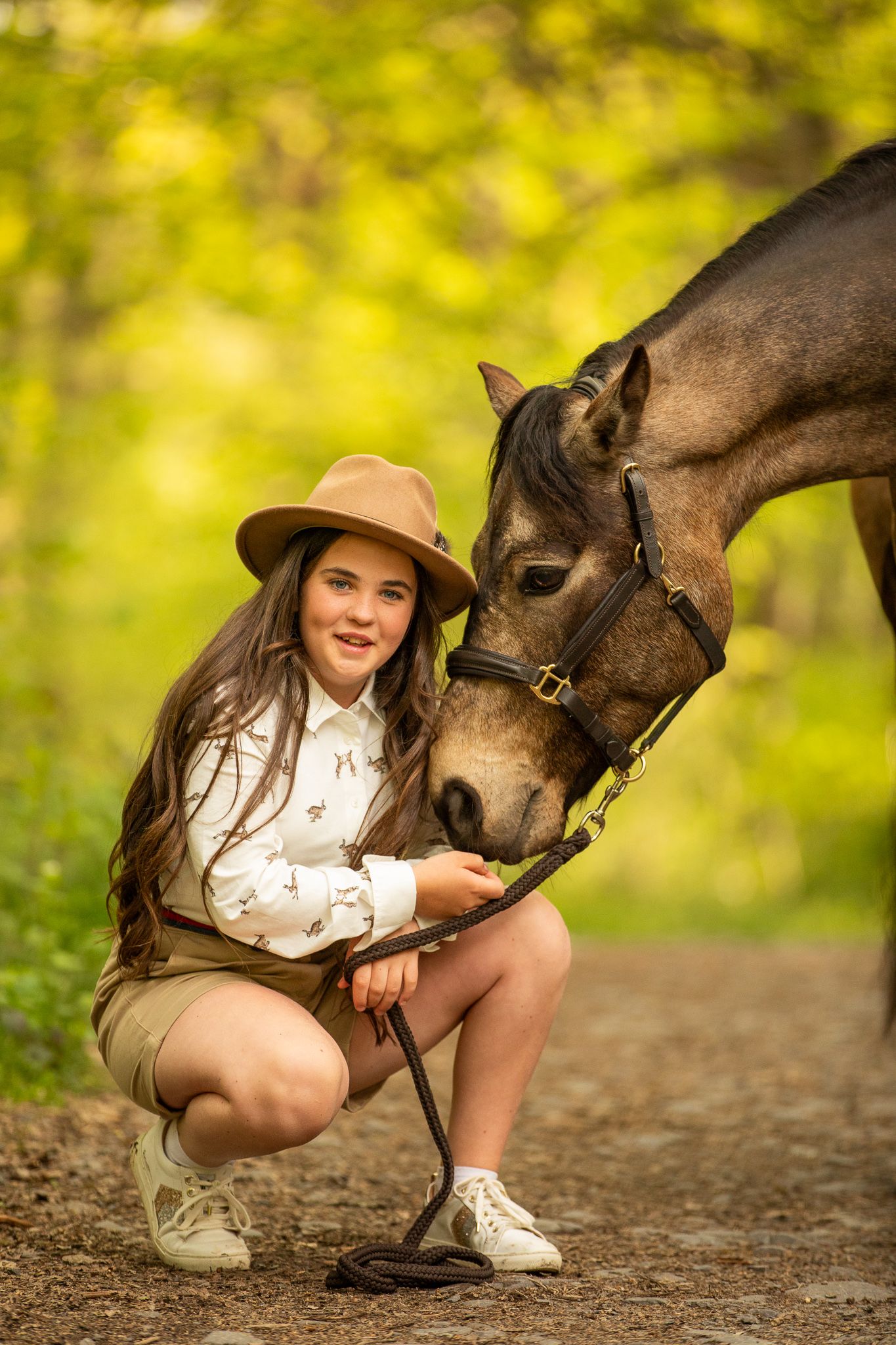 Young girl in denim overalls embraces a palomino horse in a sun-dappled forest setting.