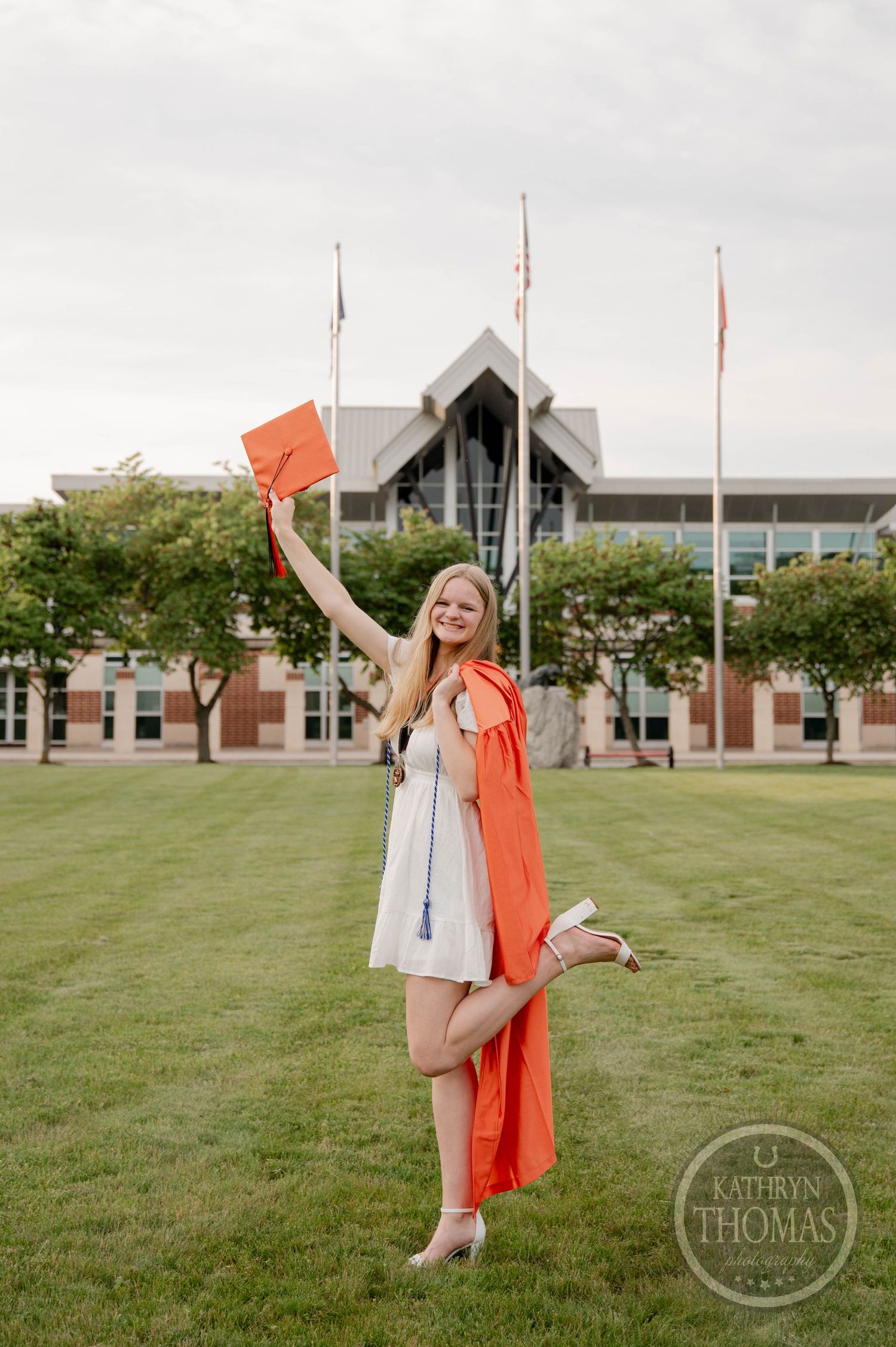A graduate in a white dress and orange graduation gown celebrates joyfully on a campus lawn with a building in the background.