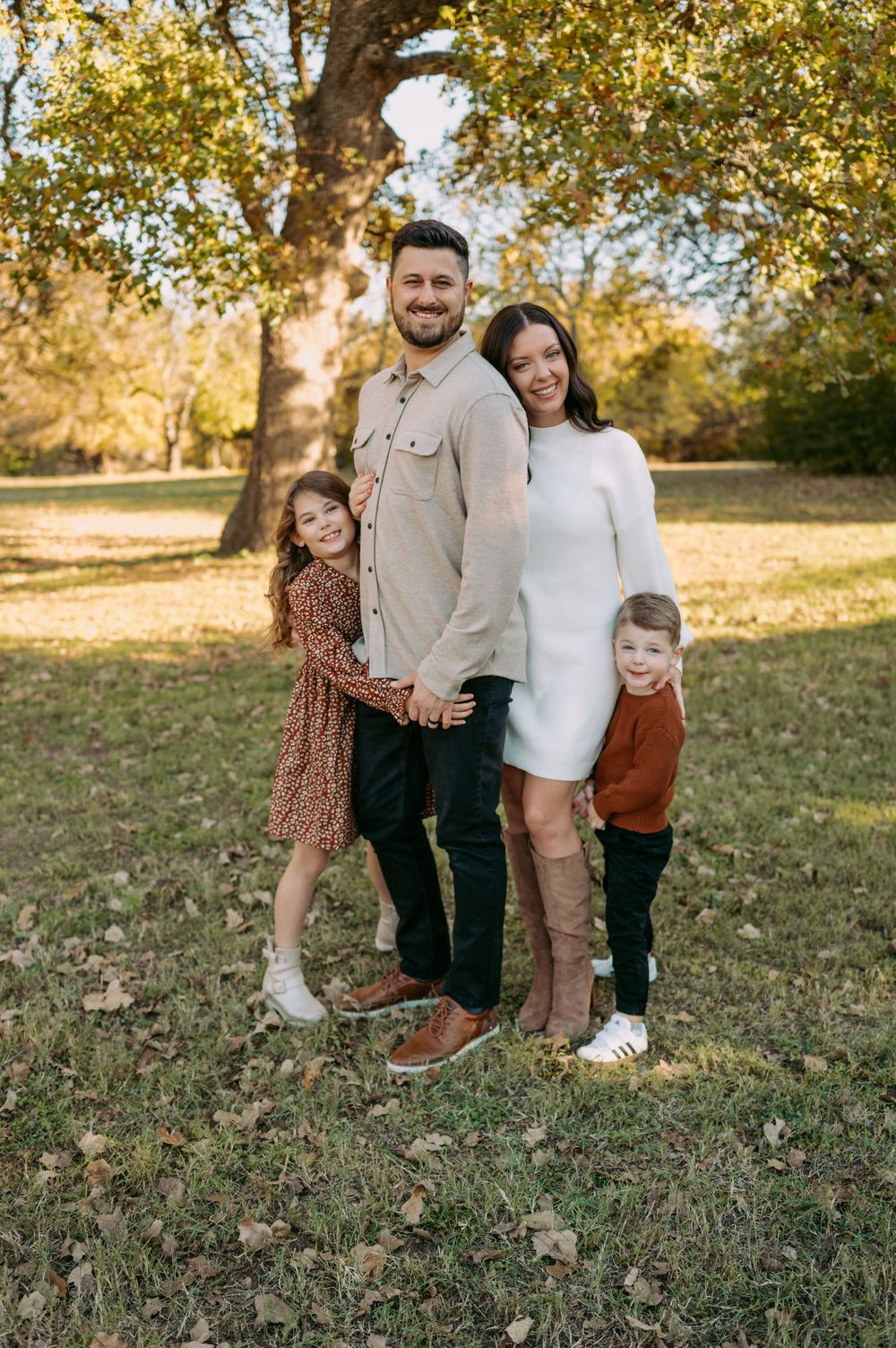 A family of four poses together outdoors in autumn, with golden leaves and sunlight filtering through trees in the background.