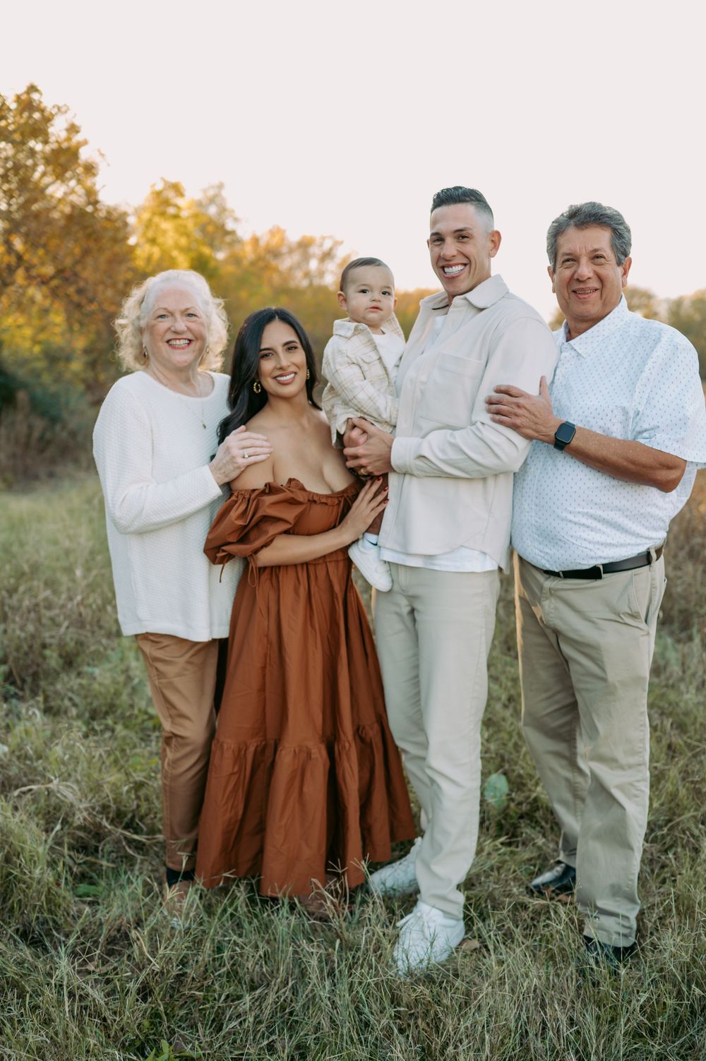 Three generations of a family pose together outdoors in a grassy field during golden hour sunset.