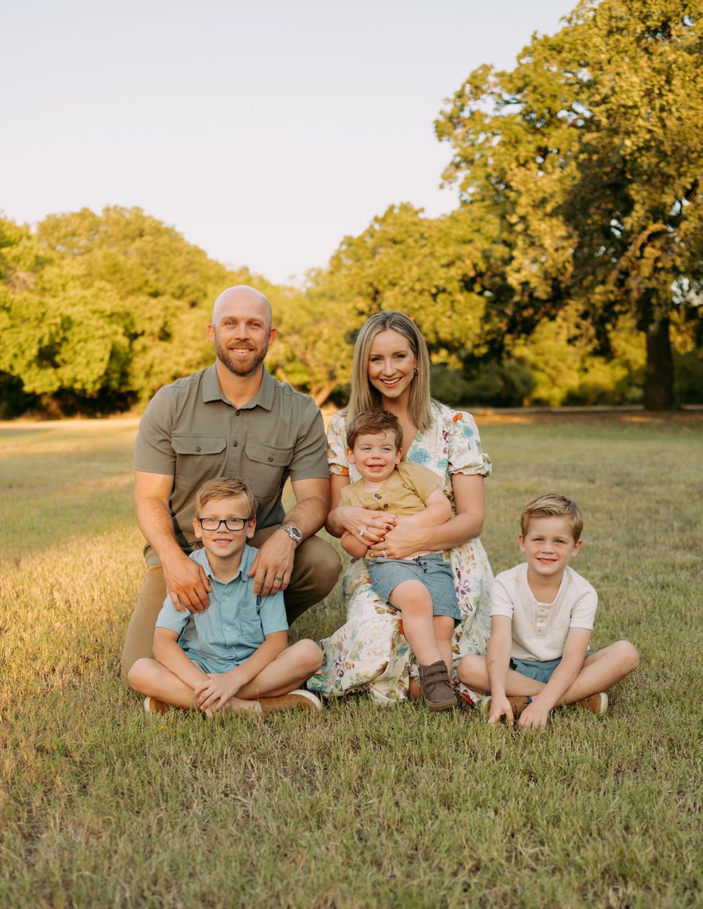 A family sits together on green grass during golden hour with autumn trees in the background.
