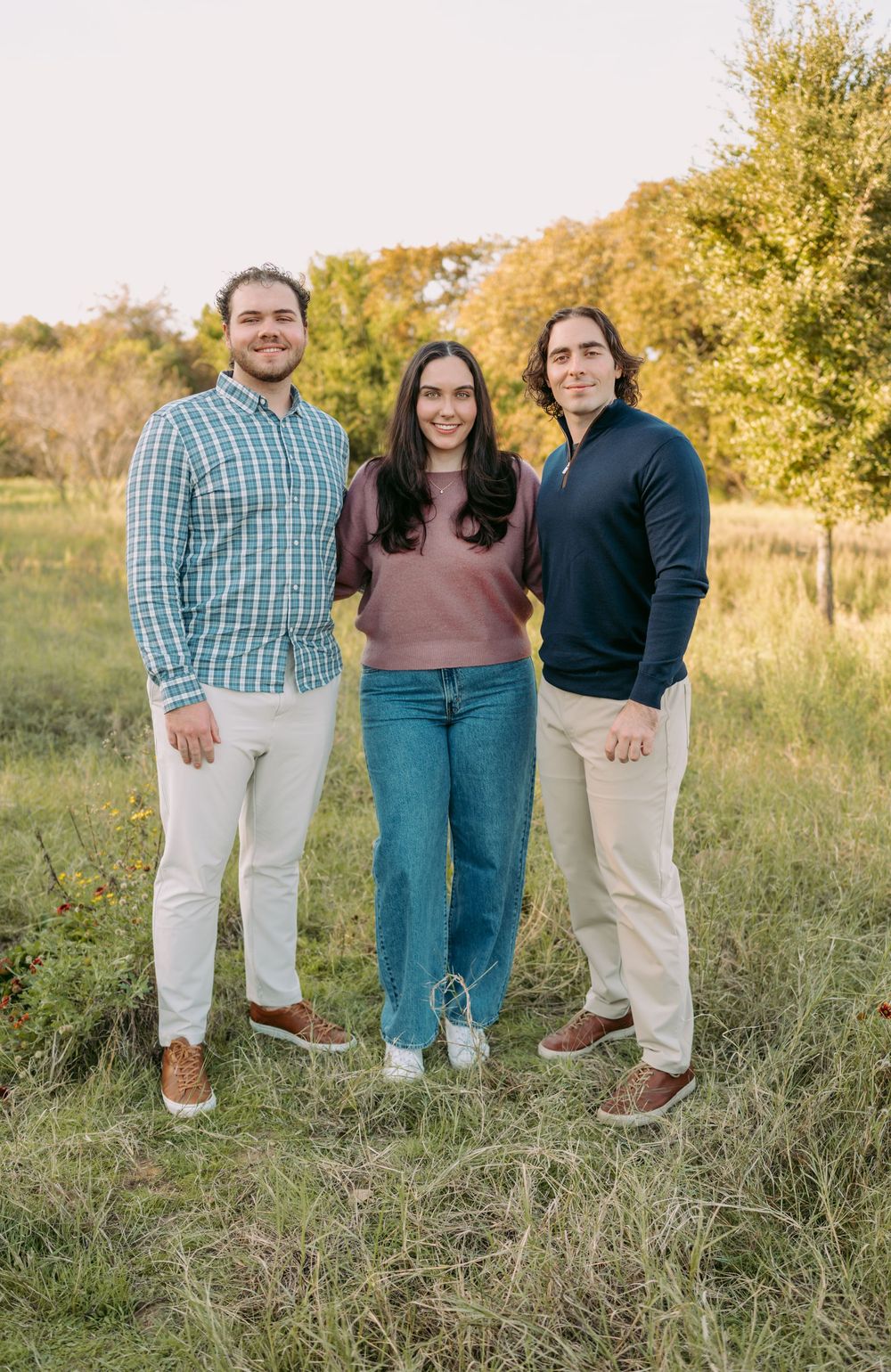 Three people stand together outdoors in casual attire against a backdrop of autumn trees and grass.