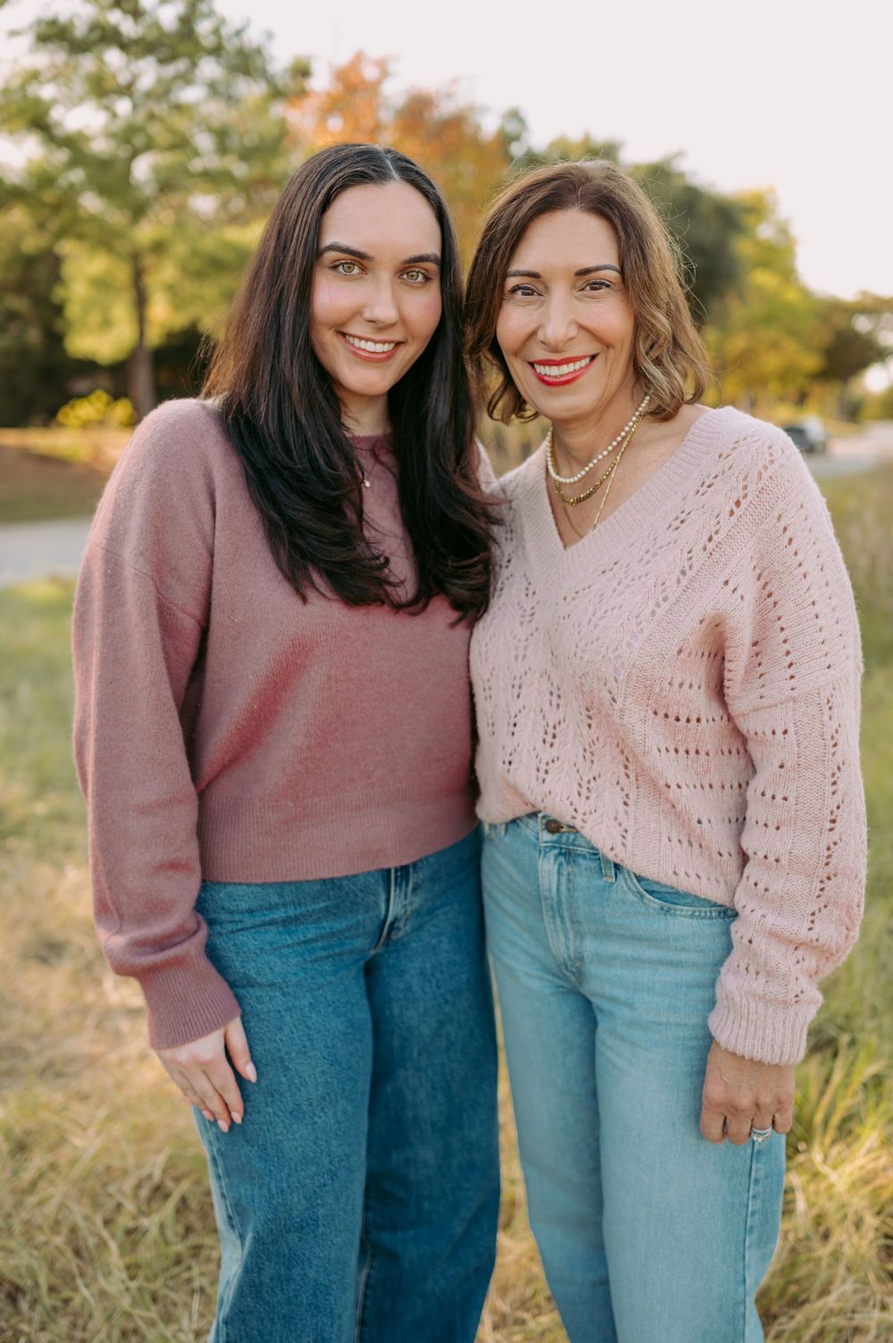 Two people wearing sweaters and jeans pose together outdoors in autumn sunlight.