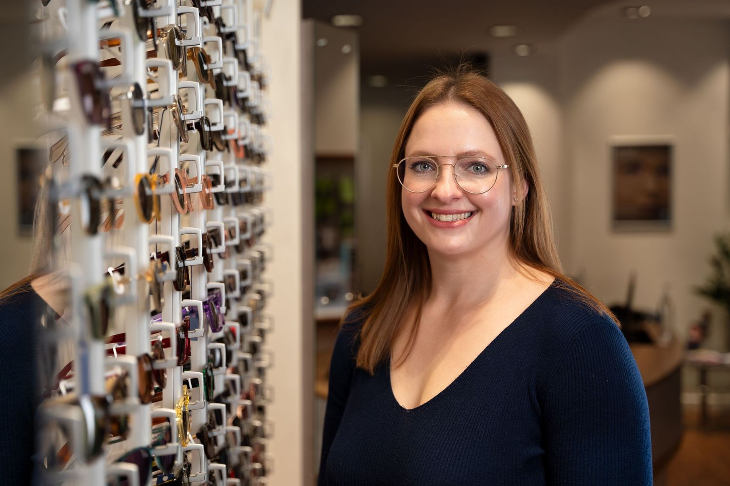 Headshot photography with a Portsmouth business. Lady stands in an Opticians with a rack of specs visible behind her.