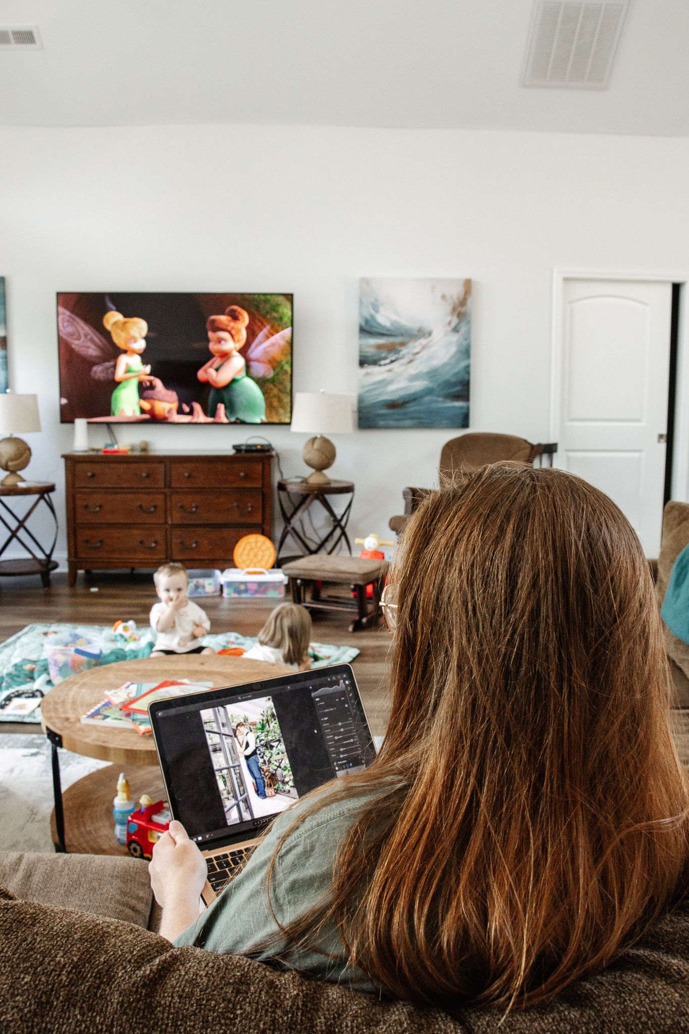 A woman is working on editing photos on her laptop, her kids in the background playing and watching a show