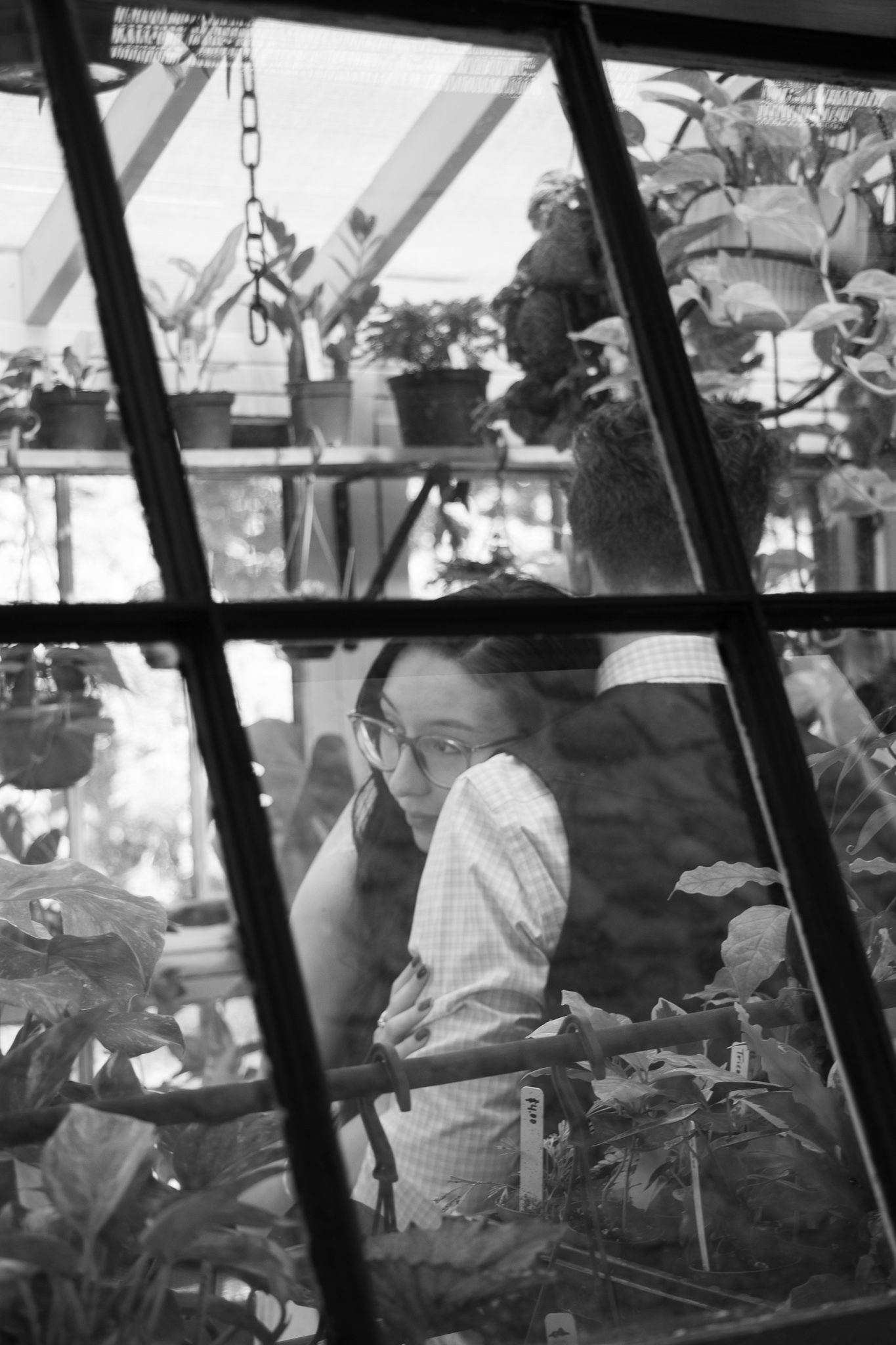 A black and white picture taken through a window focusing on bride and groom hugging inside a greenhouse.