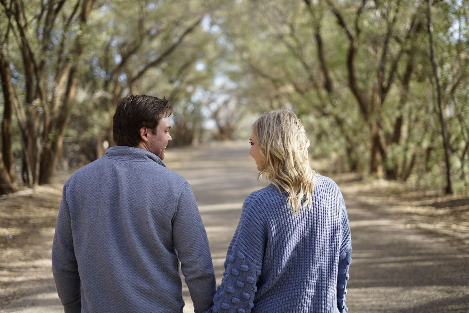 Couple walking together on tree-lined path wearing matching blue sweaters. alt="Professional  photography in Lubbock TX by H & Co. Creative