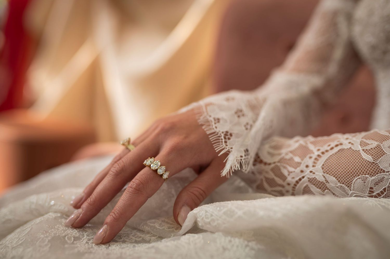 Two hands hold the delicate lace trim of a flowing white wedding dress skirt against a blush-colored background.