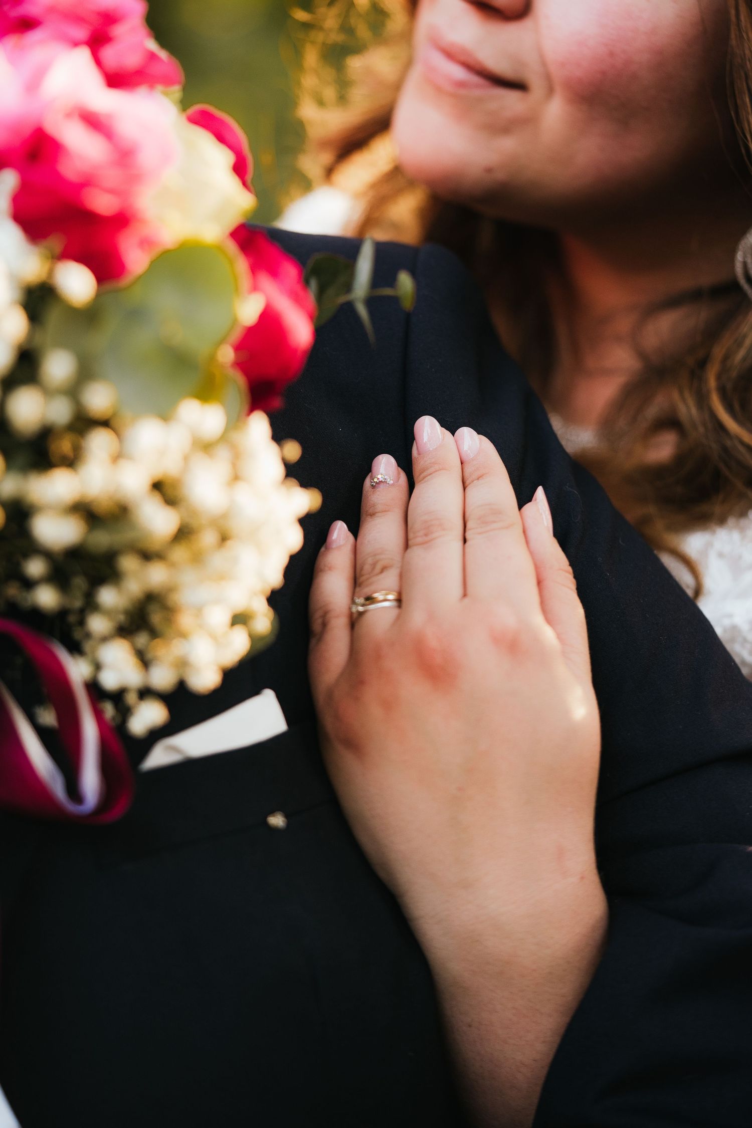 A hand wearing an engagement ring is shown against black fabric with pink flowers in soft focus.