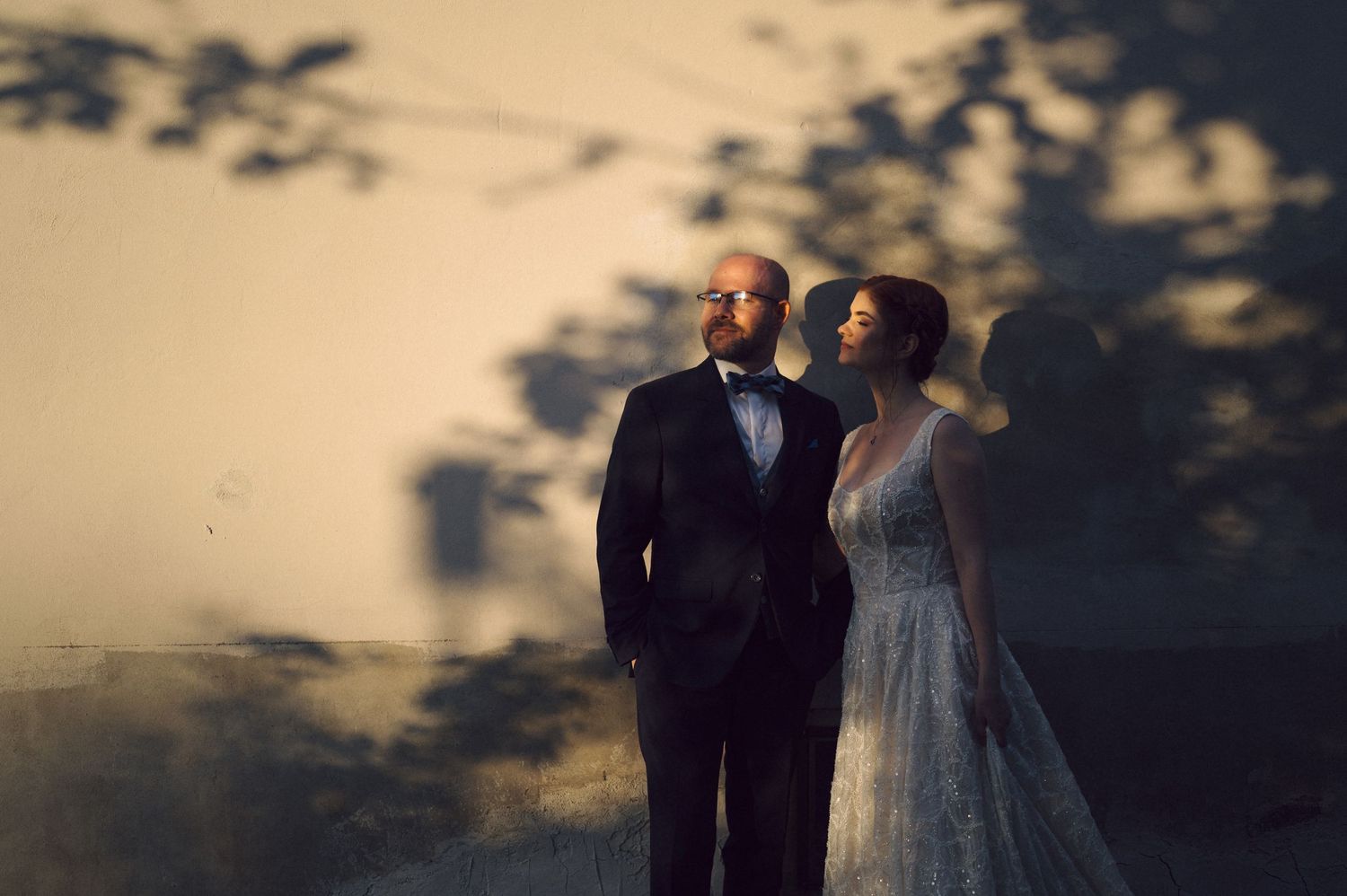 Couple in formal wedding attire standing together at dusk with dramatic tree shadow patterns on wall behind them.