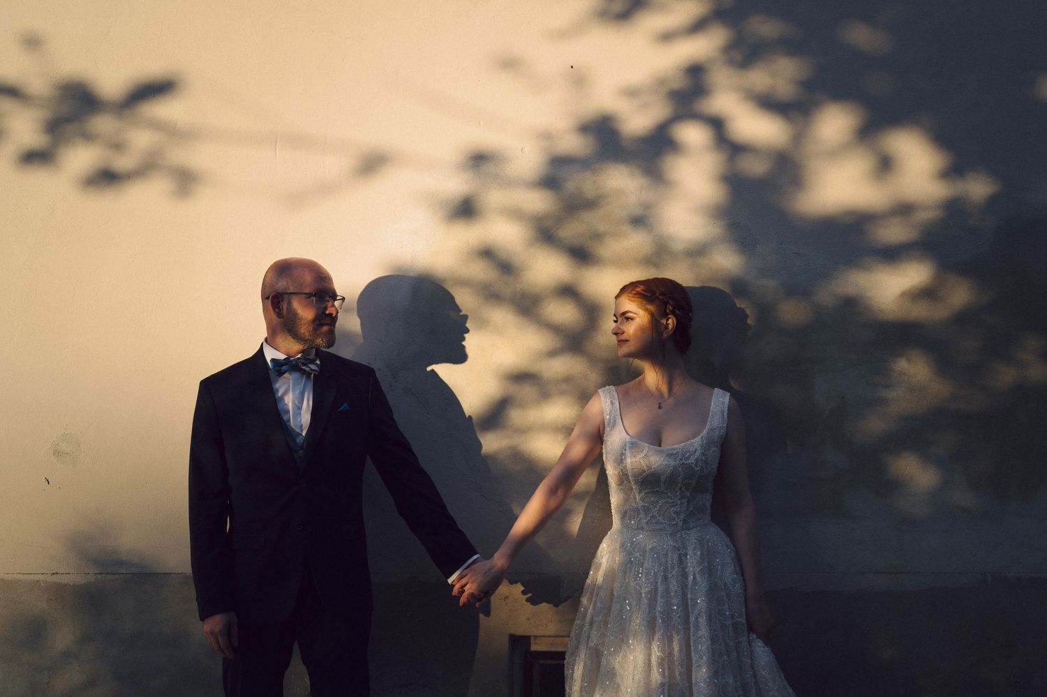 A couple in wedding attire holds hands at sunset as tree shadows dance on the wall behind them.