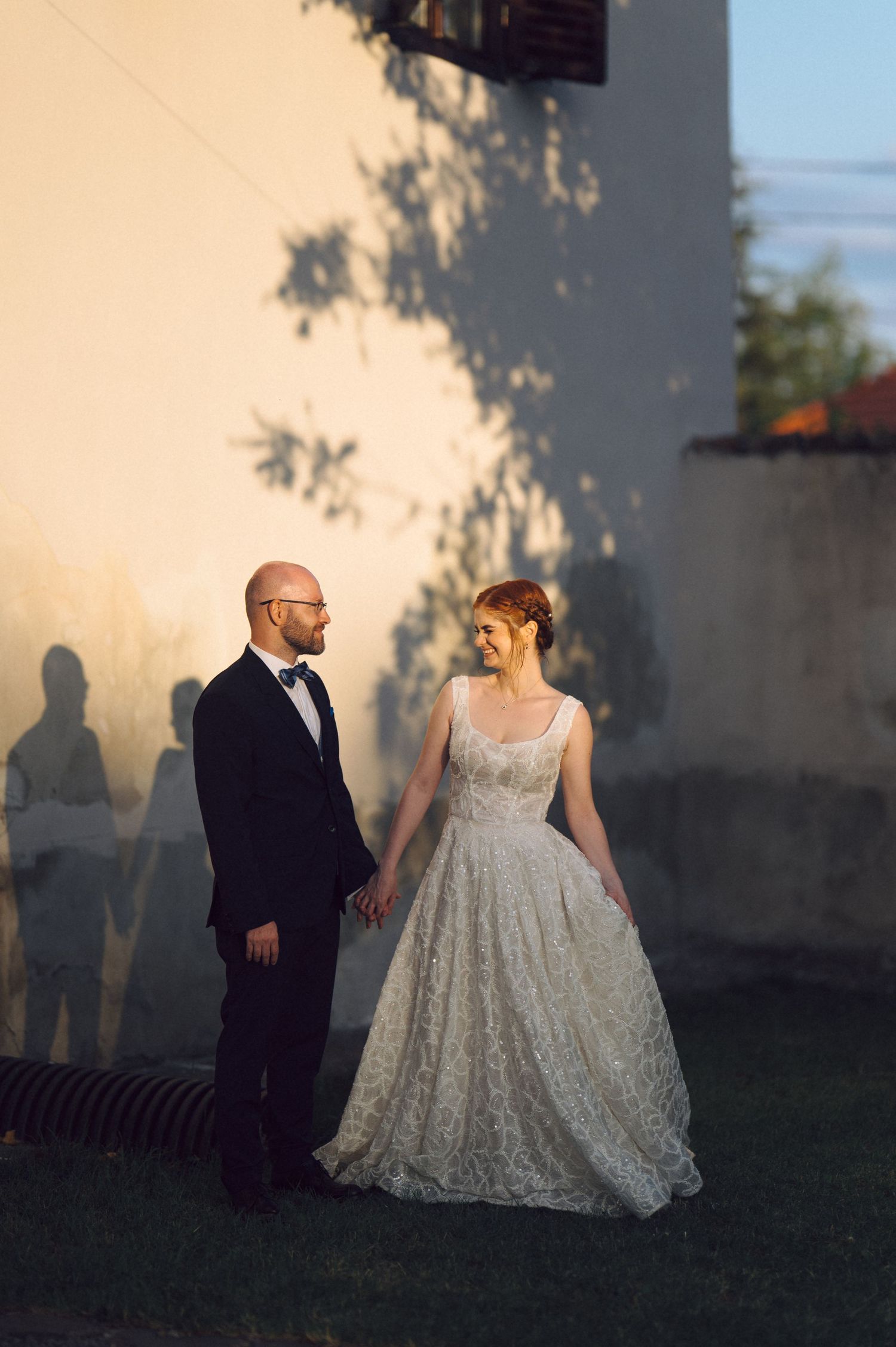 A couple stands together on a lawn at sunset, with the bride wearing a lace ball gown and the groom in a black suit.