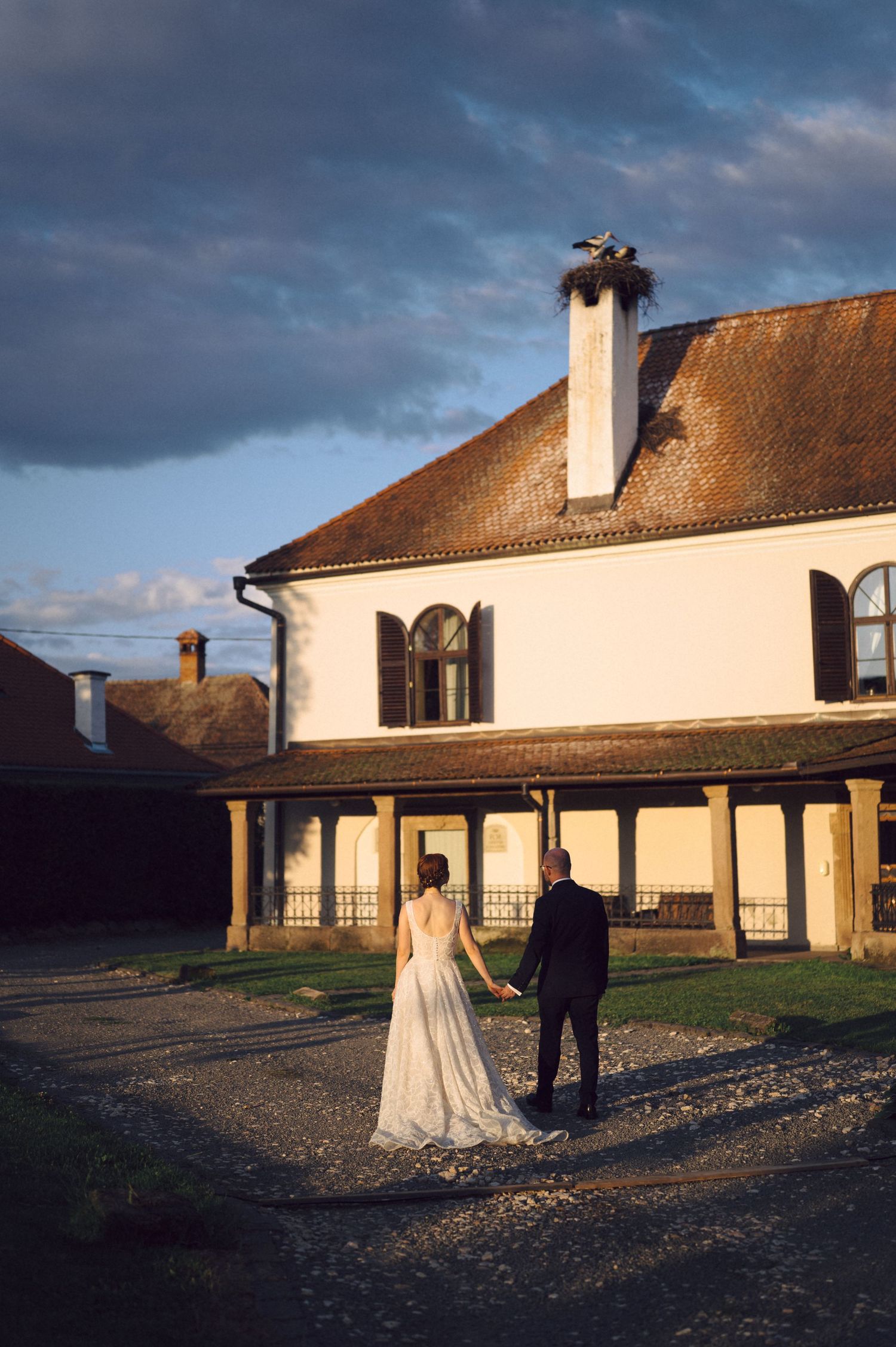 A couple in formal wedding attire stands in front of a rustic white building during sunset.